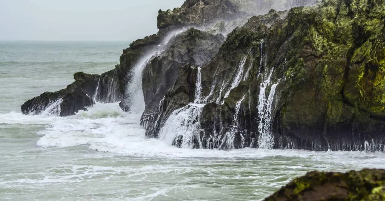 Waves crashing against a rocky, waterfall-lined coastline where a river meets the ocean