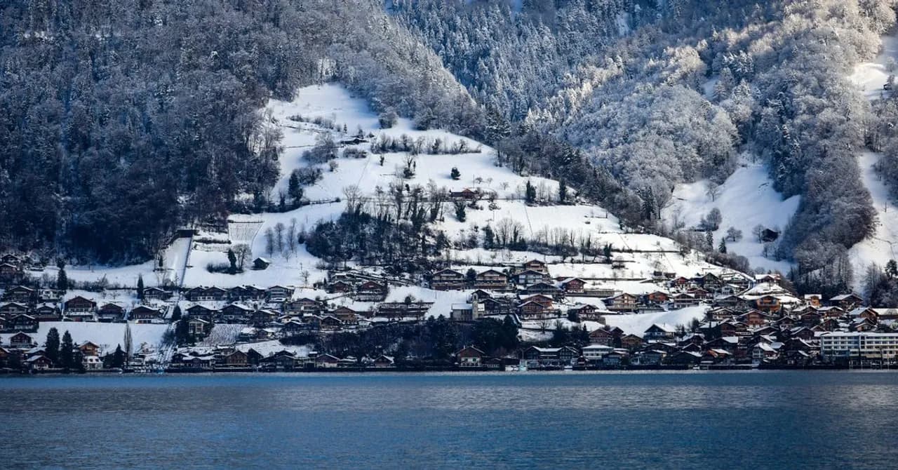 A Swiss village of wooden chalets along a lake shore beneath snow-covered alpine forest