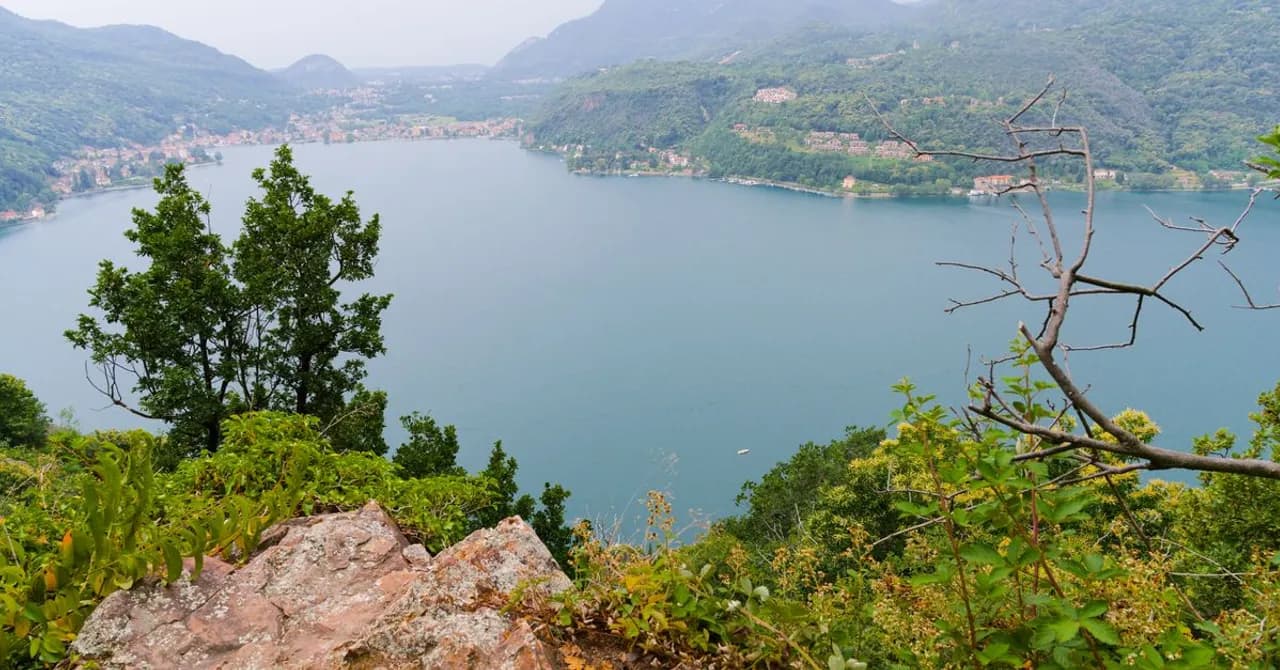 Ein See, umgeben von steilen bewaldeten Hügeln unter dunstigem Alpenhimmel