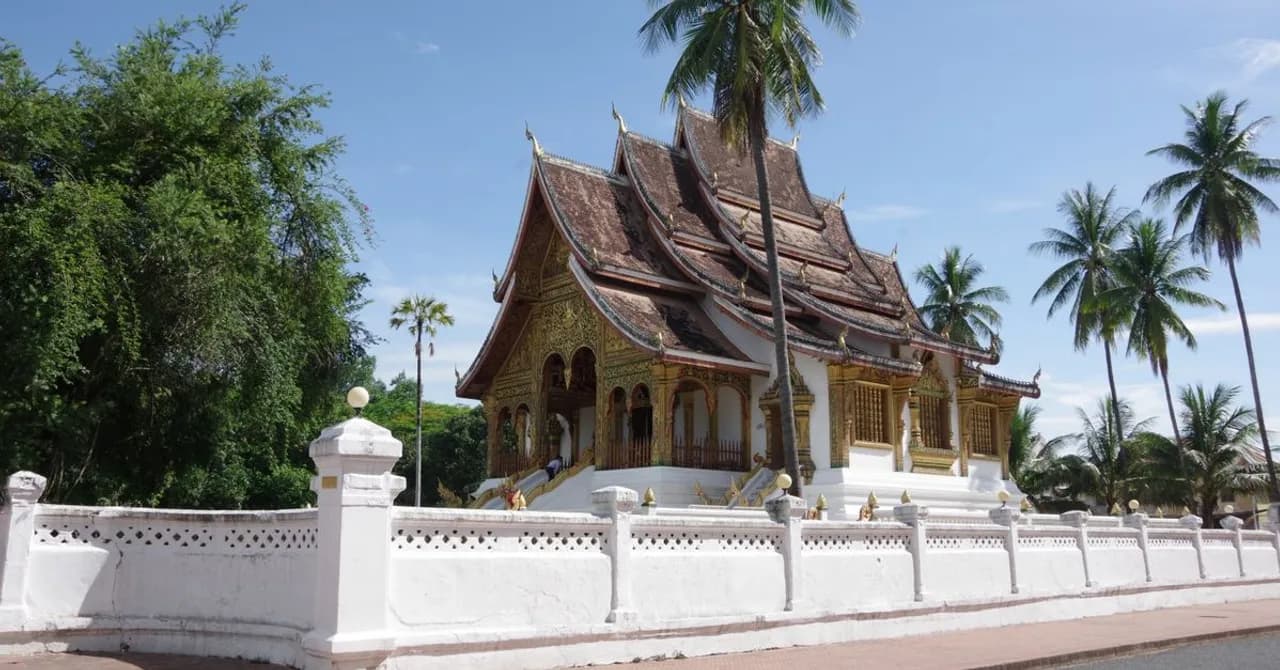 Un tempio laotiano dal tetto dorato a più falde dietro un muro bianco e alcune palme