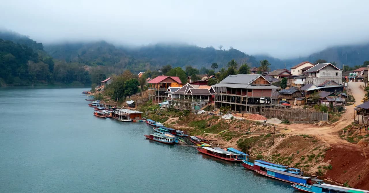 Barche di legno ormeggiate sotto un villaggio sul fiume, con le montagne nebbiose del Laos settentrionale