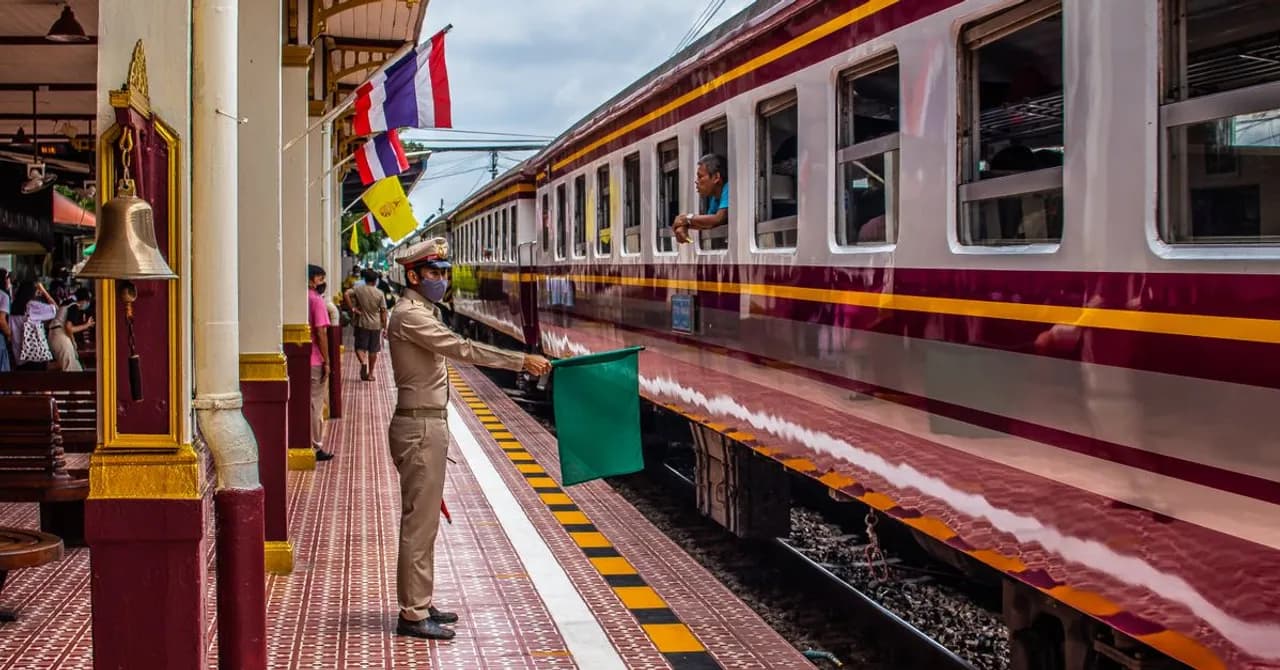 Ein Zug wartet an einem Bahnsteig in Thailand, daneben Personal mit Signalfahne